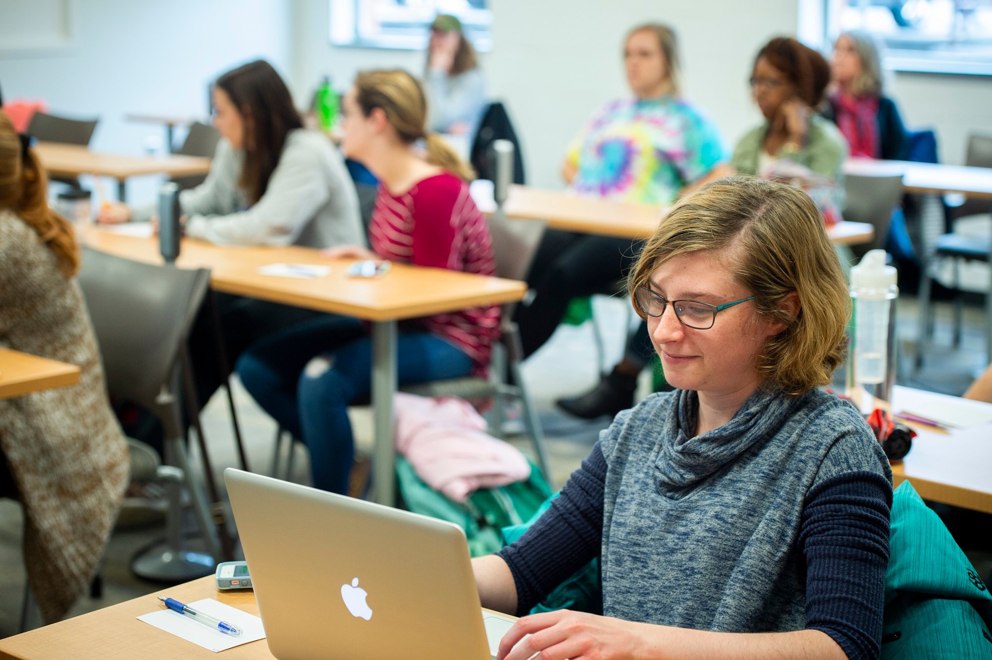 A light-skinned female student working on a laptop in a crowded classroom.