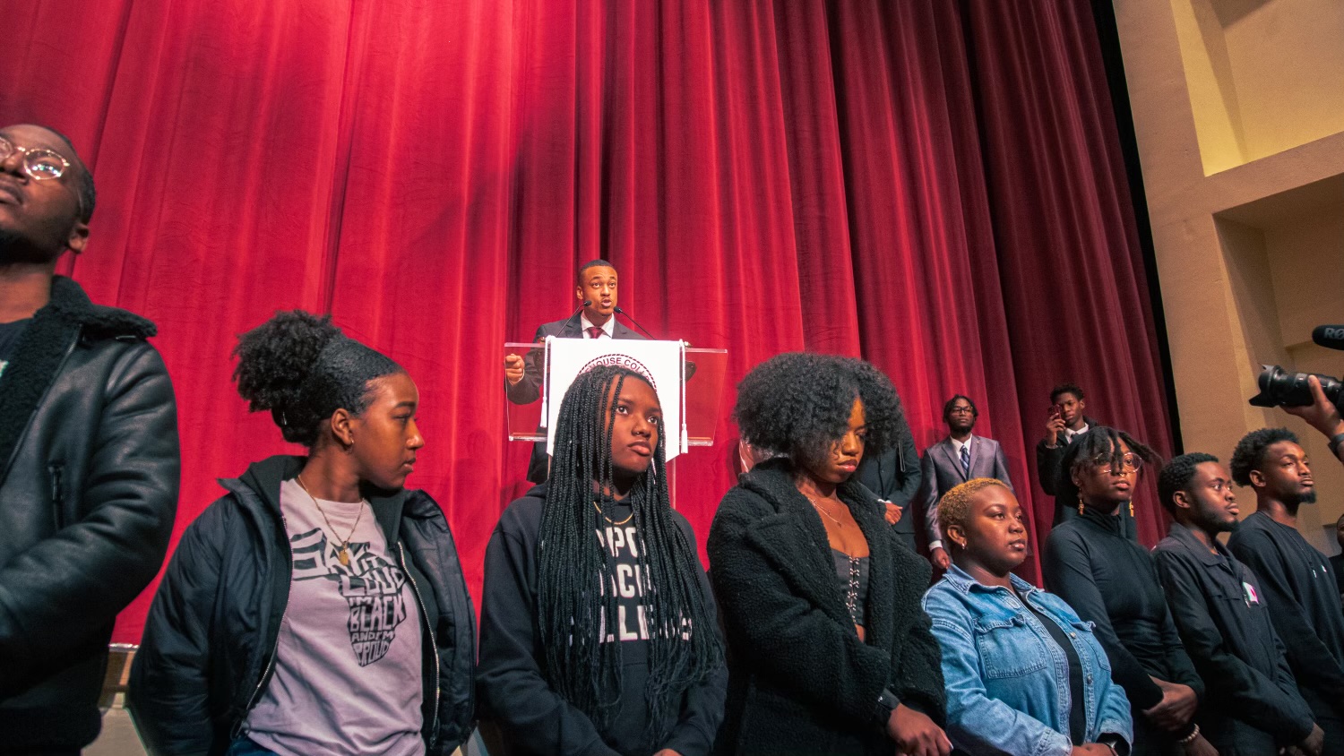 Student Daxton Pettus speaks at a podium with a red velvet curtain behind him and students standing in front of him in solidarity.