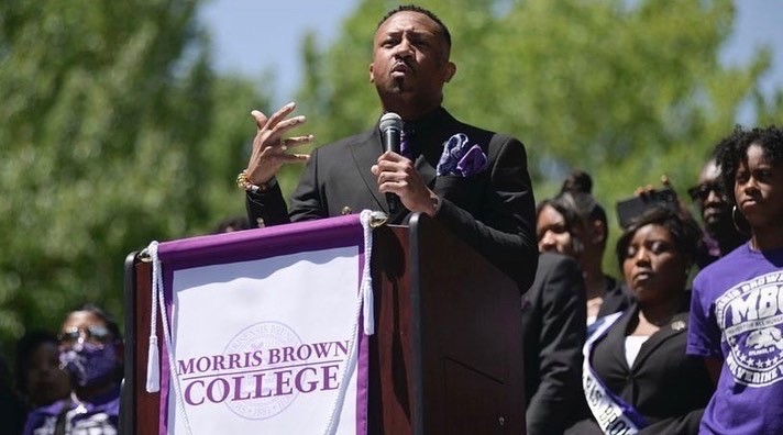 Morris Brown College president Kevin James stands at a podium with a microphone. Students in purple college spirit wear stand in the background.