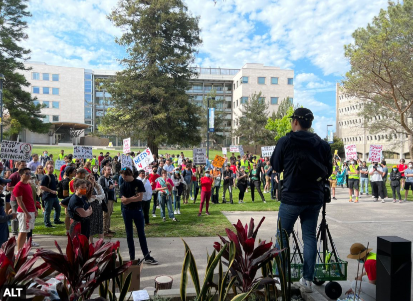 A person in the foreground speaks at a podium. A crowd of people is assembled.