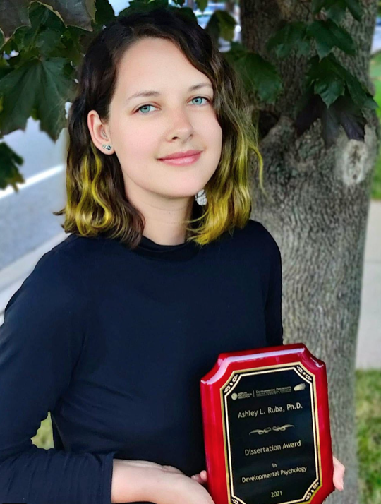 Ashley Ruba, a light-skinned woman with wavy hair, holds her award from the APA.