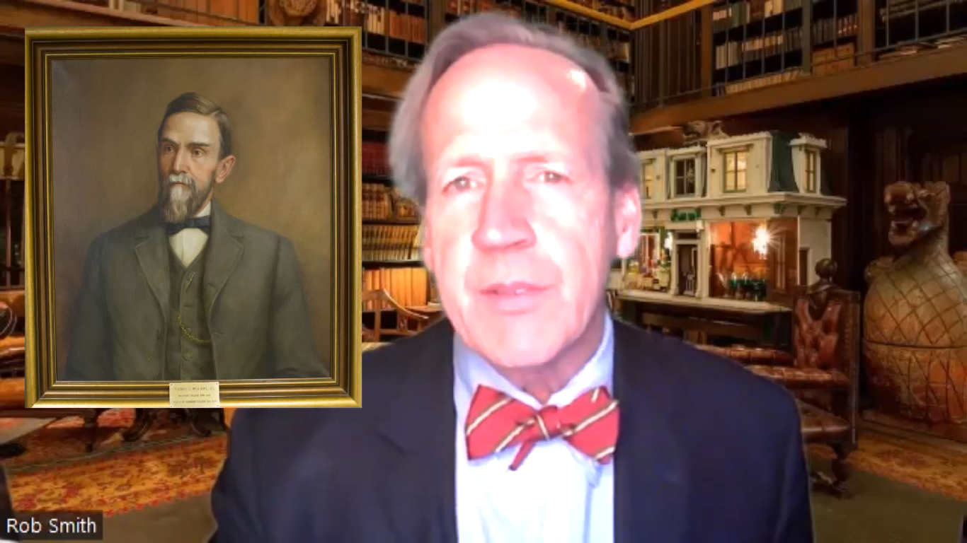 a man in a bowtie talks next to a portrait of an older man from the 1800s 