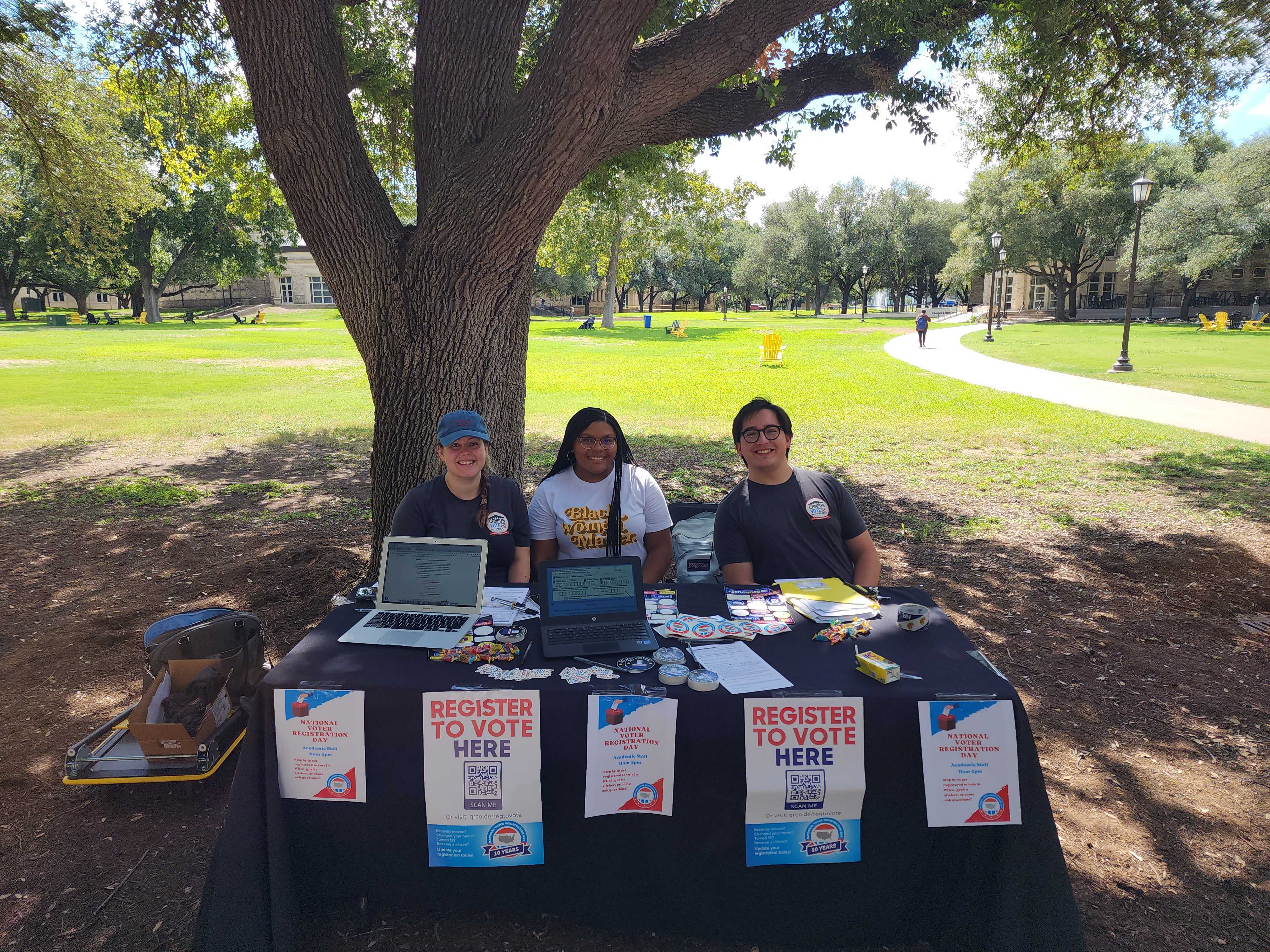Three students sit at a table in front of a tree on a green lawn. The sign on the table reads "register to vote here."