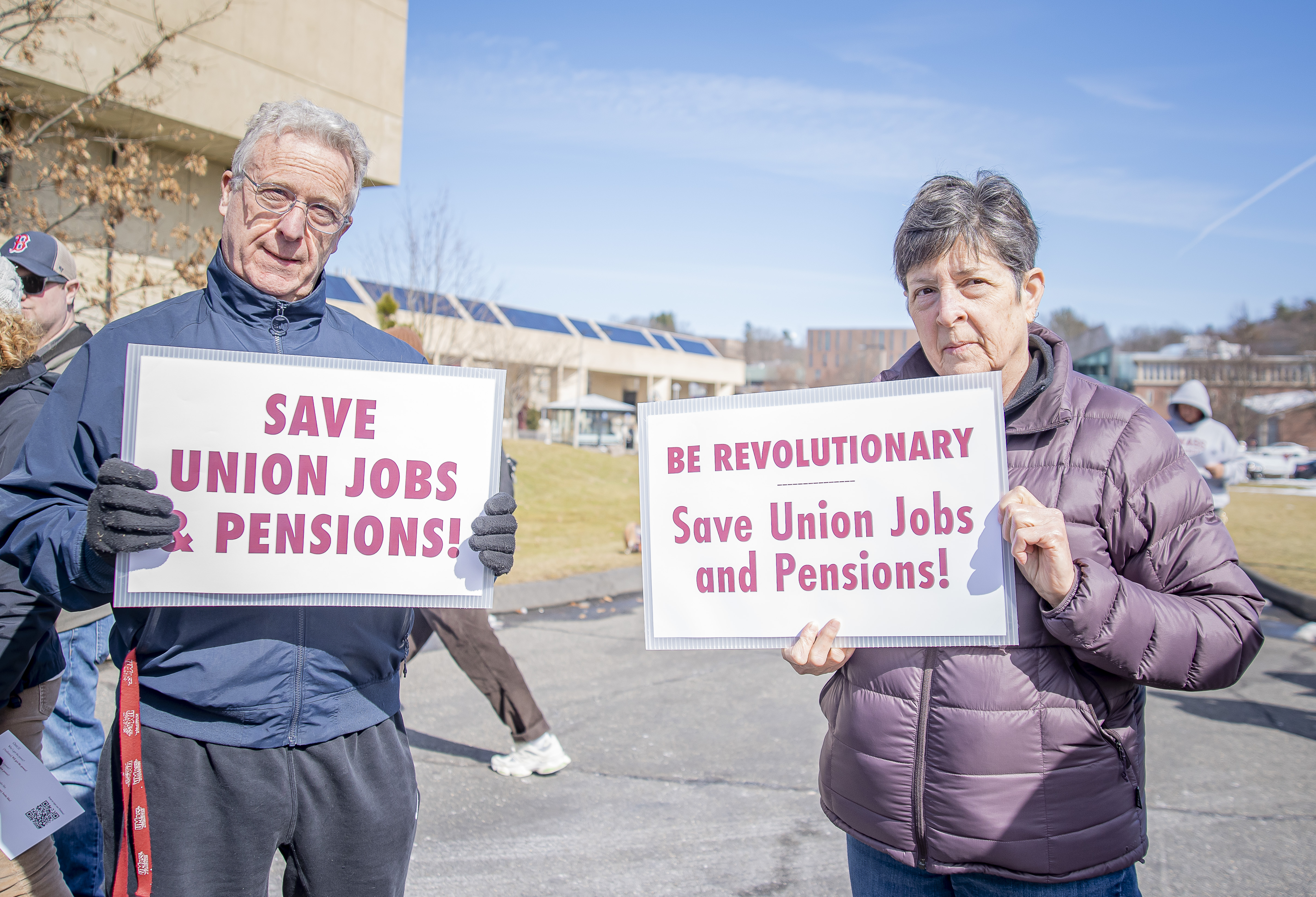 An older man and a woman stand side by side holding signs that say, "save union jobs & pensions."