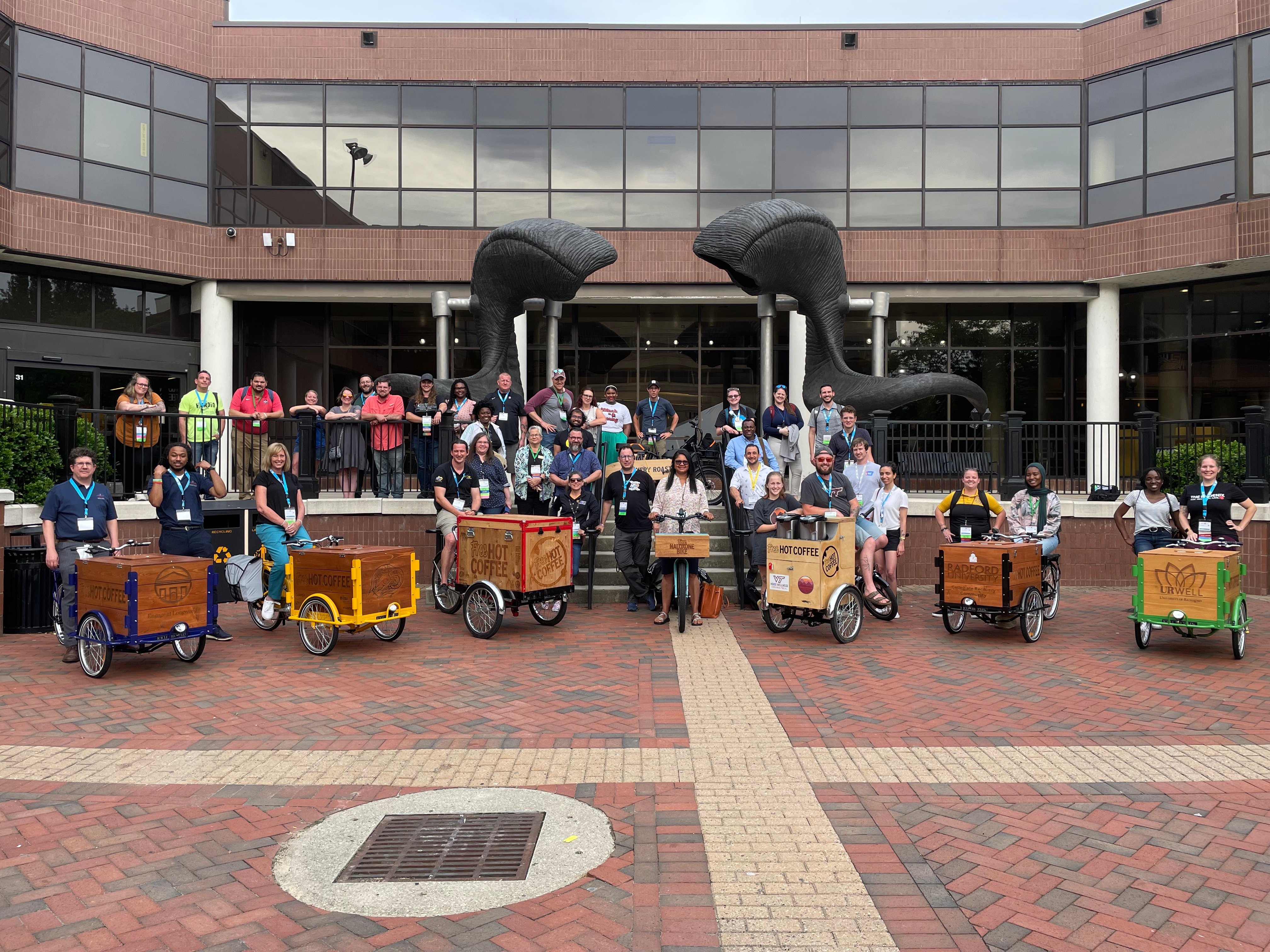 Leaders of campus recovery centers are gathered on the steps of a building with giant ram's horn statue above them and carts of coffee in front.