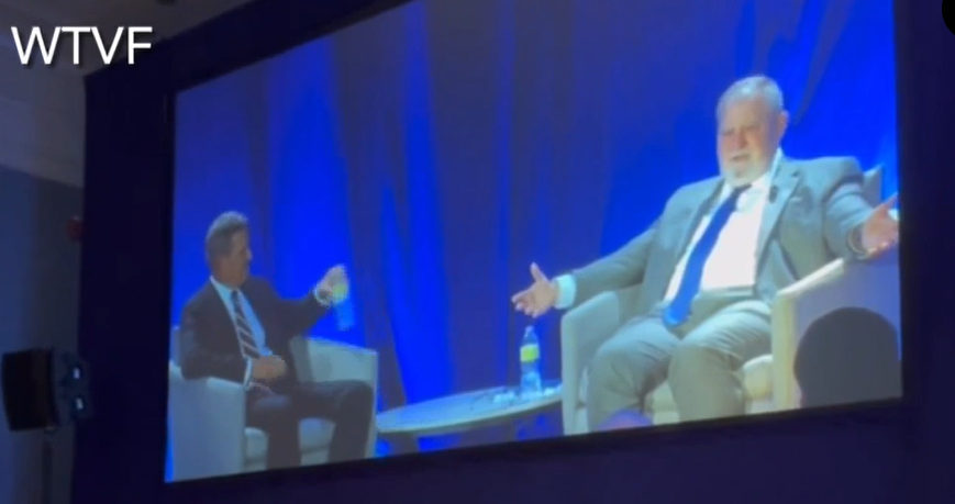 Photo of a video screen showing two white men seated in armchairs on a stage.