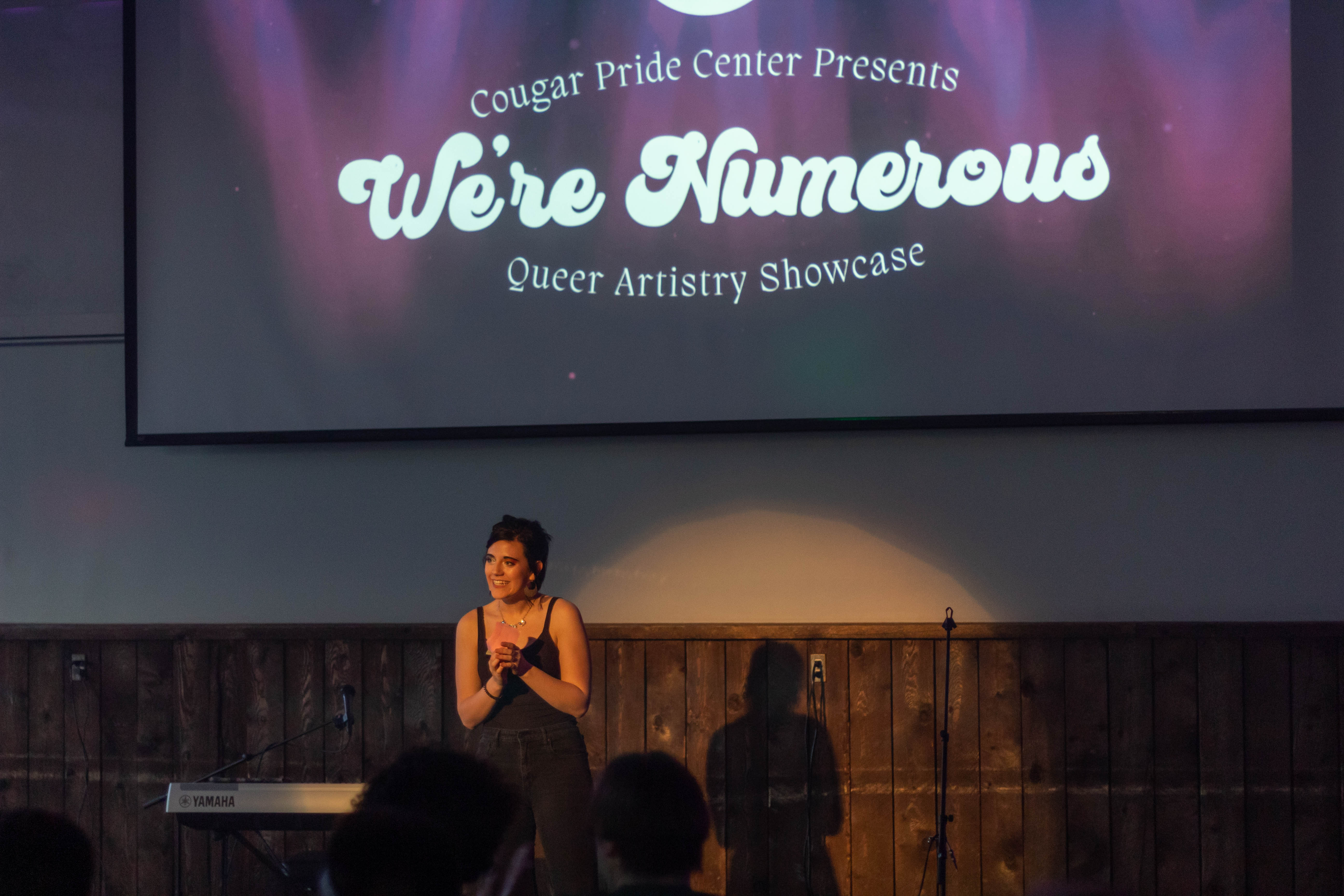 A woman stands on a stage under a screen showing the words "Cougar Pride Center Presents 'We're Numerous' Queer Artistry Showcase."