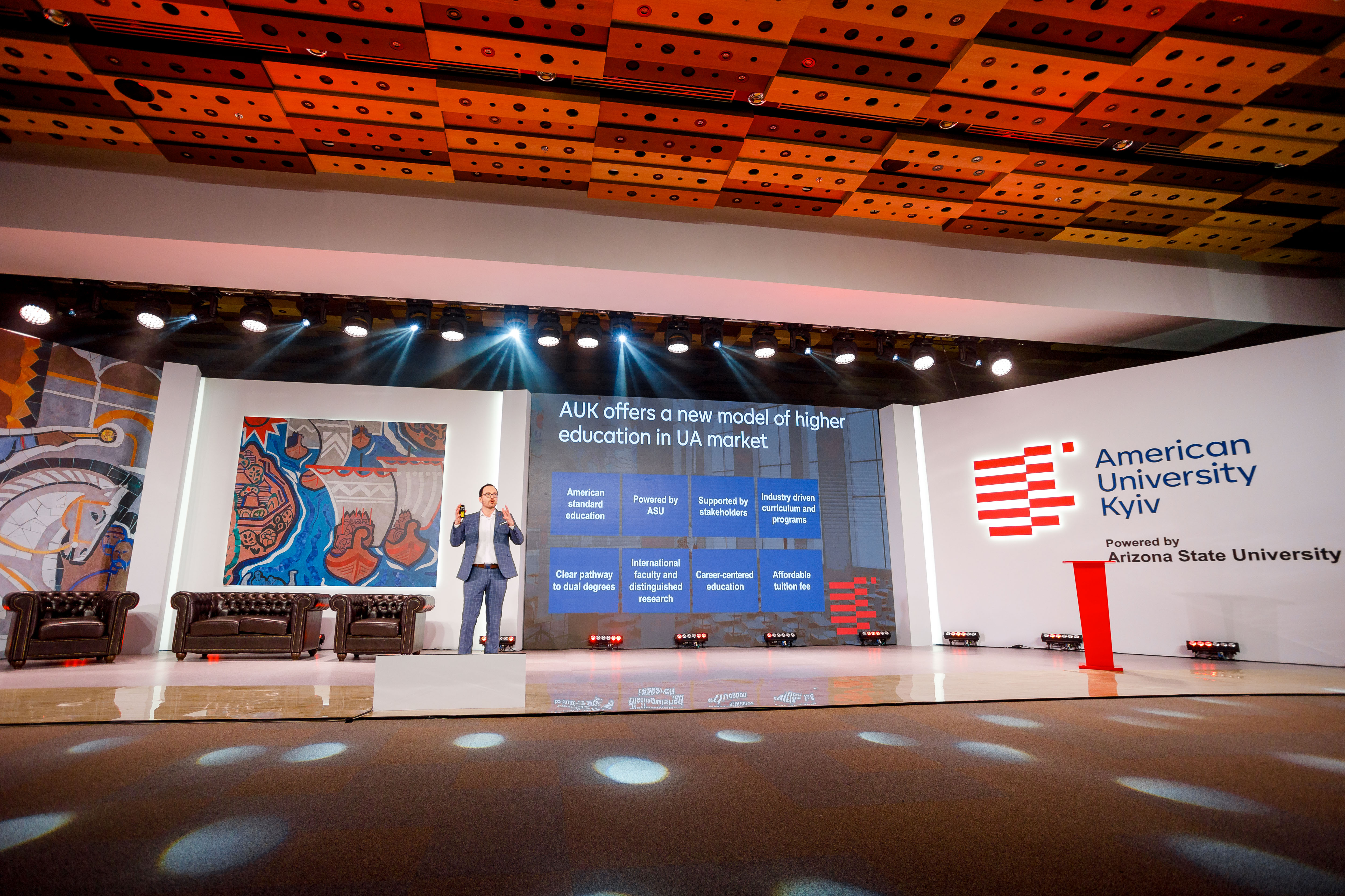 A man in a suit stands on stage in front of a projected slide and talks