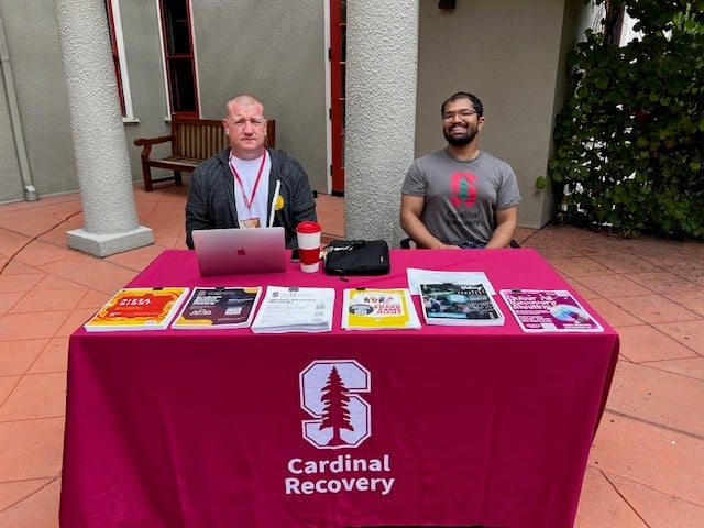 Two men sit behind a burgundy table with fliers on top at Stanford University.