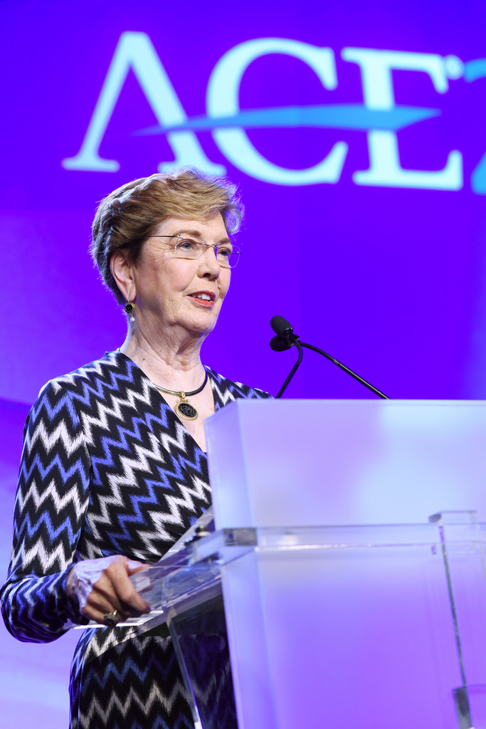 Molly Broad, a white woman with short hair and glasses wearing a dress with a zigzag pattern, stands in front of a purple screen showing the American Council on Education logo.