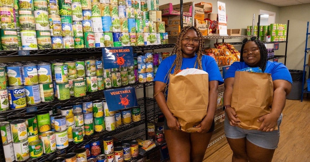 Two dark-skinned women wearing blue shirts hold bags of groceries and stand in front of shelves of canned food.