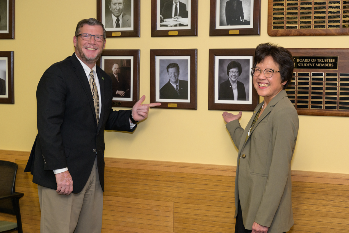Tom Ramage, a white man, and Pamela Lau, an Asian woman, point at the official portraits.