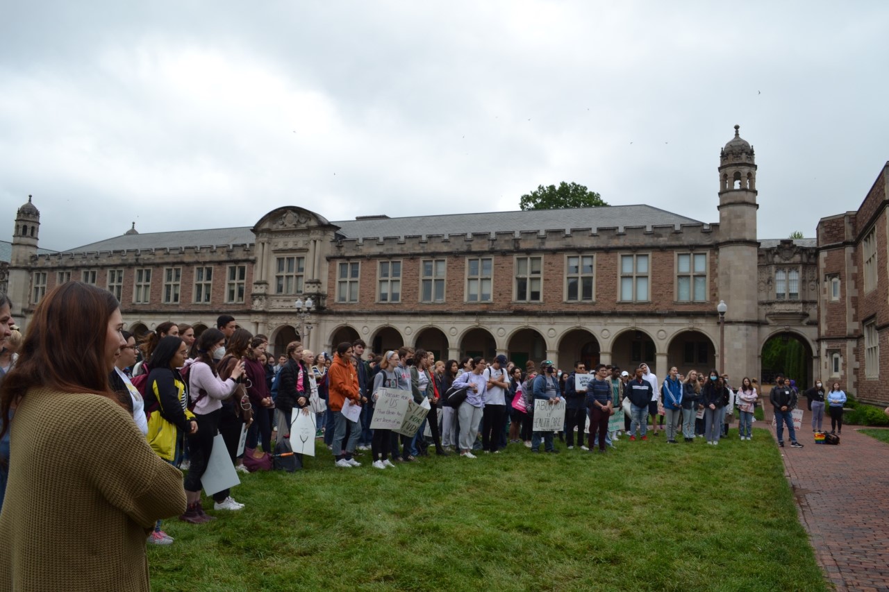 Students are assembled on a lawn in front of an academic building. Many carry signs.